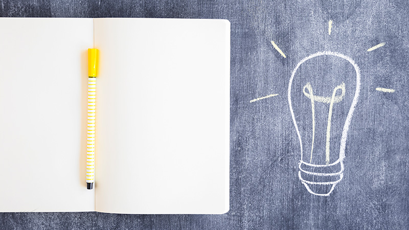 open notebook with a pen next to a drawing of a light bulb on a chalkboard